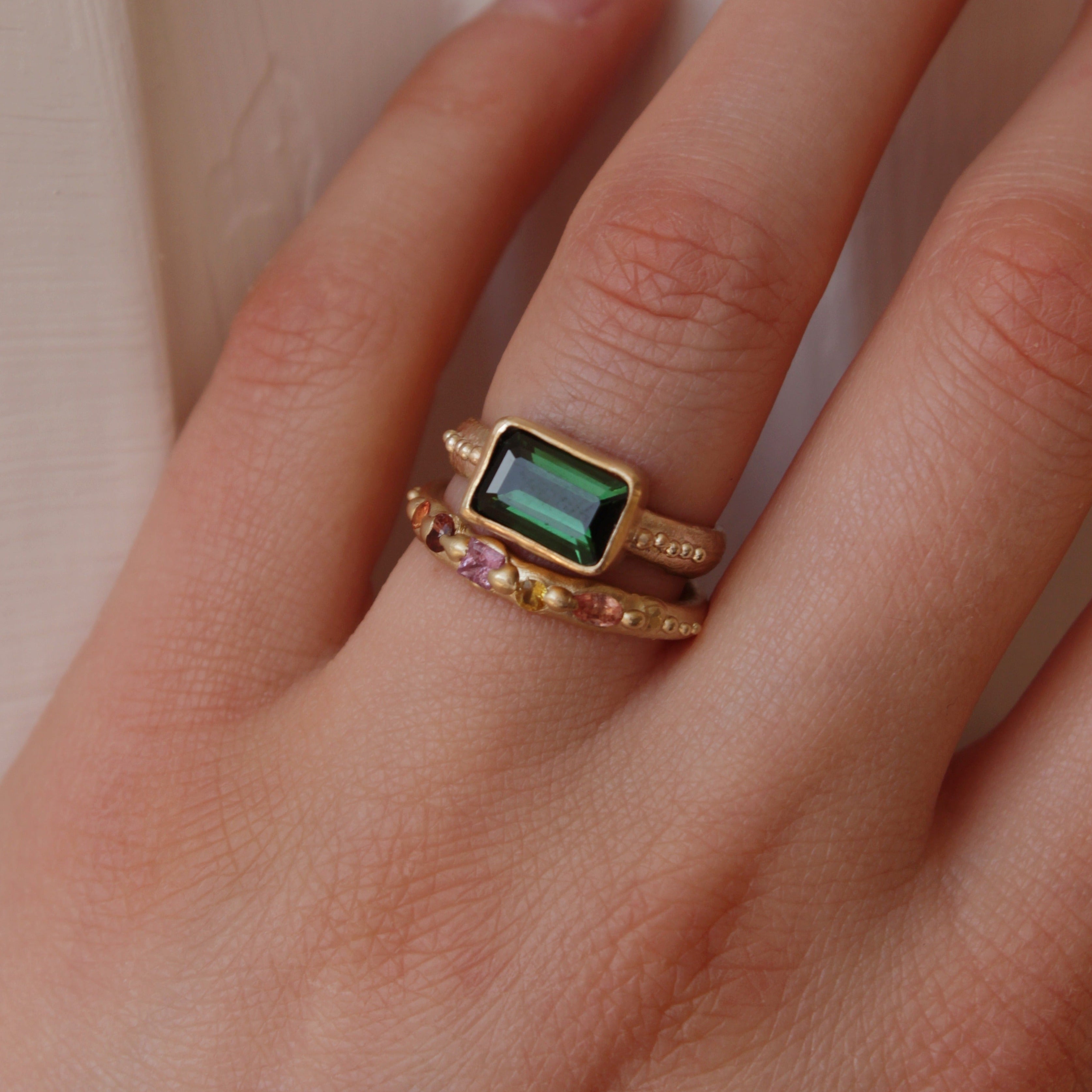 A stunning Green Tourmaline stone set on a 9 ct gold band. The band features an organic textures with tinny gold granules delicately framing the centrepiece stone.. Hand crafted alternative engagement ring by Josie Mitchell jewellery. Made in Frome, Somerset. Worn with the Sapphire wedding ring in 9 ct gold.